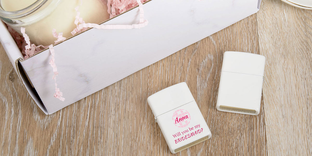 Two personalized cream-colored lighter-shaped keepsakes on a light wood surface, with one showing a pink printed message and the other closed. A white marble-patterned gift box with pink shredded paper is partially visible in the background.