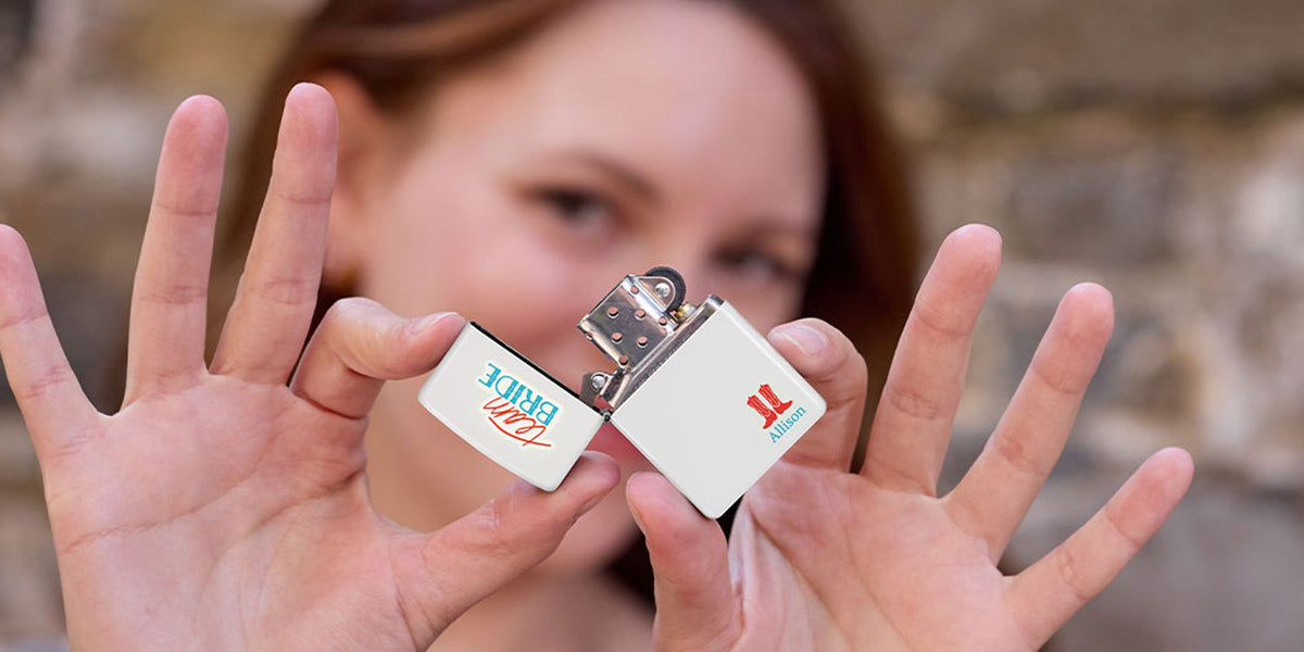 A close-up lifestyle shot of a woman holding a small white lighter with printed logos, held up by her hands against a blurred stone wall background.
