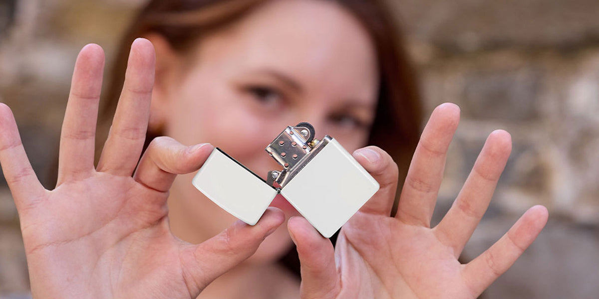 A close-up lifestyle shot of a woman holding a white matte Zippo lighter, held up by her hands against a blurred stone wall background.