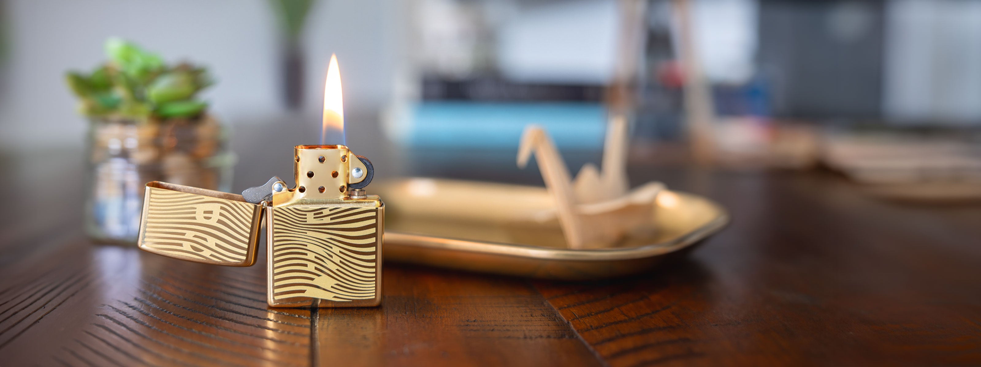 A gold-toned lighter with a wavy striped pattern is shown lit on a wooden table, with a blurred decorative rolling tray and small plant in the background.