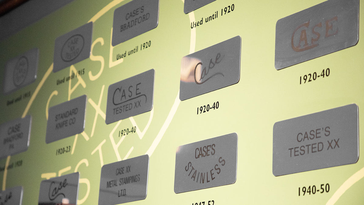A close-up of a vintage display wall featuring multiple gray rectangular plaques with engraved text such as 'CASE', 'CASE'S BRADFORD', 'CASE'S STAINLESS', and dates like '1920-40' and '1940-50' on a pale yellow background.