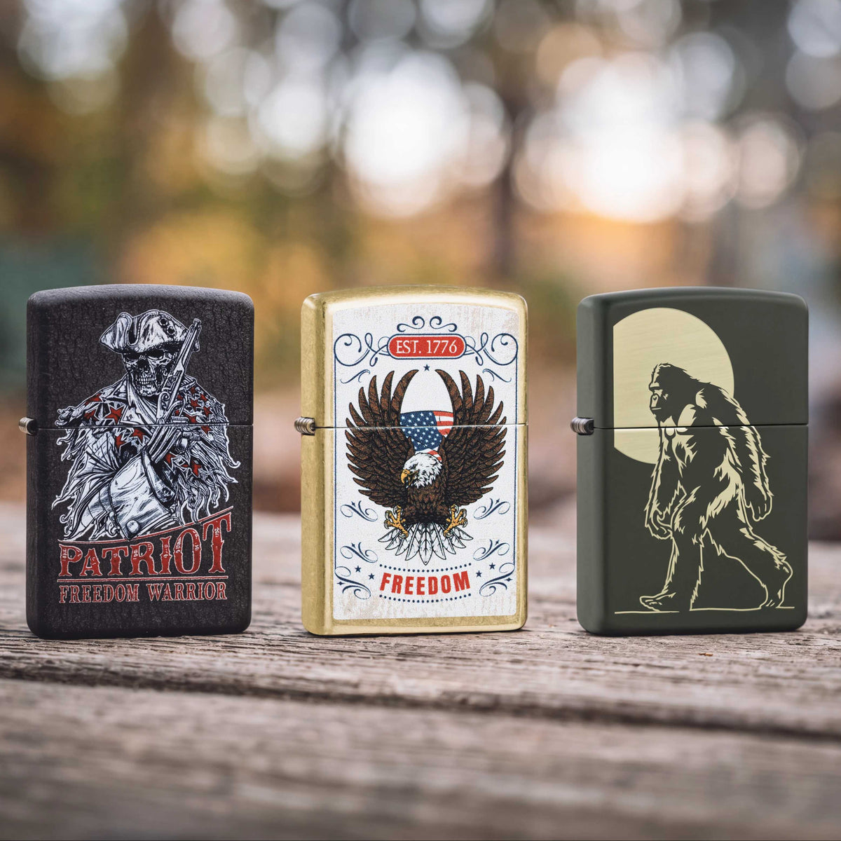 Three Zippo lighters on a wooden surface. First one says "Patriot Freedom Warrier', the middle one is an eagle on a white background, and the third is a Sasquatch.