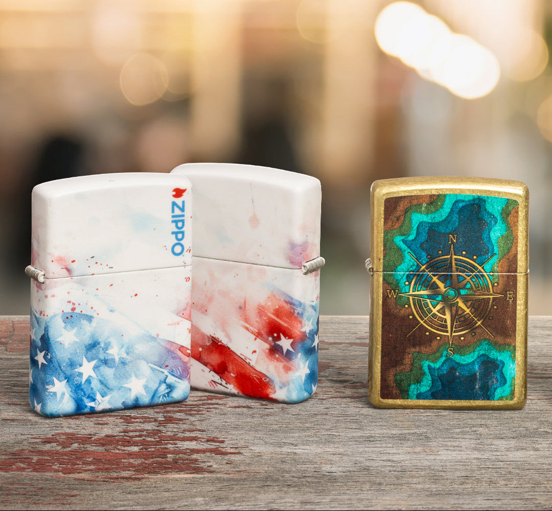 Three Zippo lighters, two with colorful designs and the other with a compass rose design, on a wooden surface.
