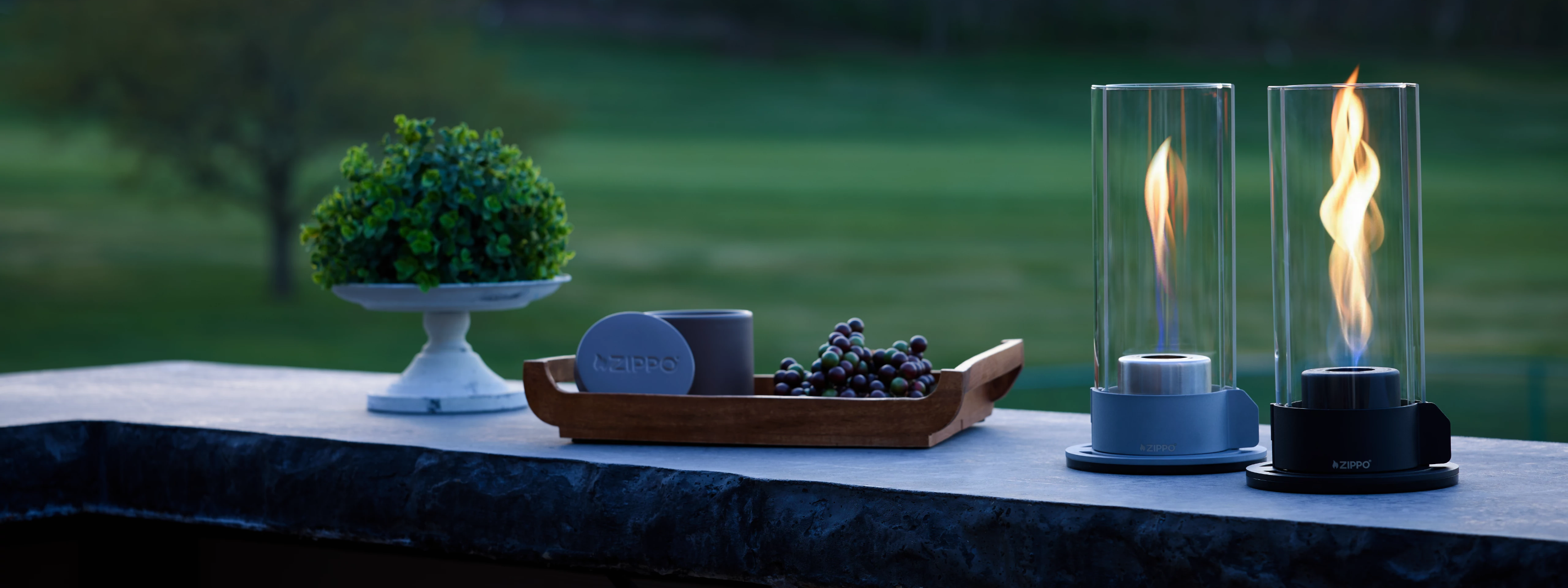 Two Zippo Flamescapes and a Citronella candle in an outdoor space with grass in the background.