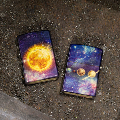 Lifestyle image of two Zippo Solar Flare Design 540 Tumbled Brass Windproof Lighters lying on the ground; one showing the front of the lighter while the other shows the back of the lighter. 

