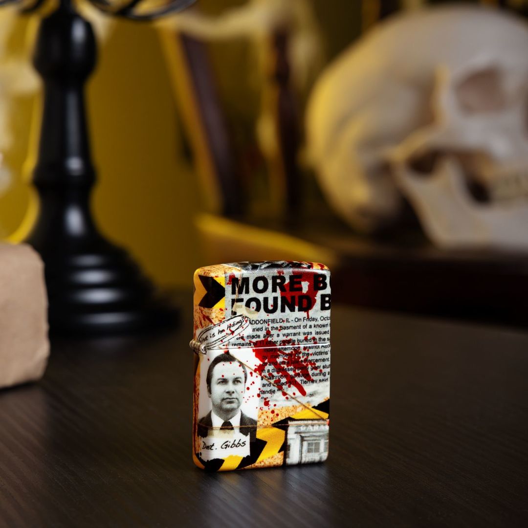 Lifestyle image of Zippo Murder Mystery Design 540 Matte Windproof Lighter standing on a table in front of a black candelabra showing the front of the lighter with a creepy background with a skull on a chair. 

