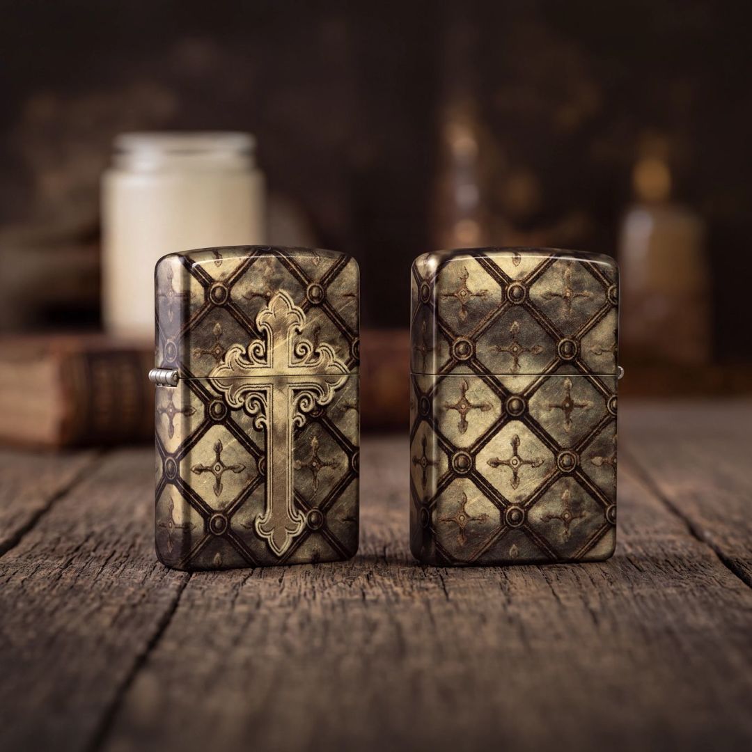 Lifestyle image of two Zippo Gilded Cross Design 540 Tumbled Brass Windproof Lighters both standing on a wooden surface one showing the front and hinge side of the lighter while the other is showing the back and non-hinge side of the lighter. The background is out of focus with jar candles and a book. 

