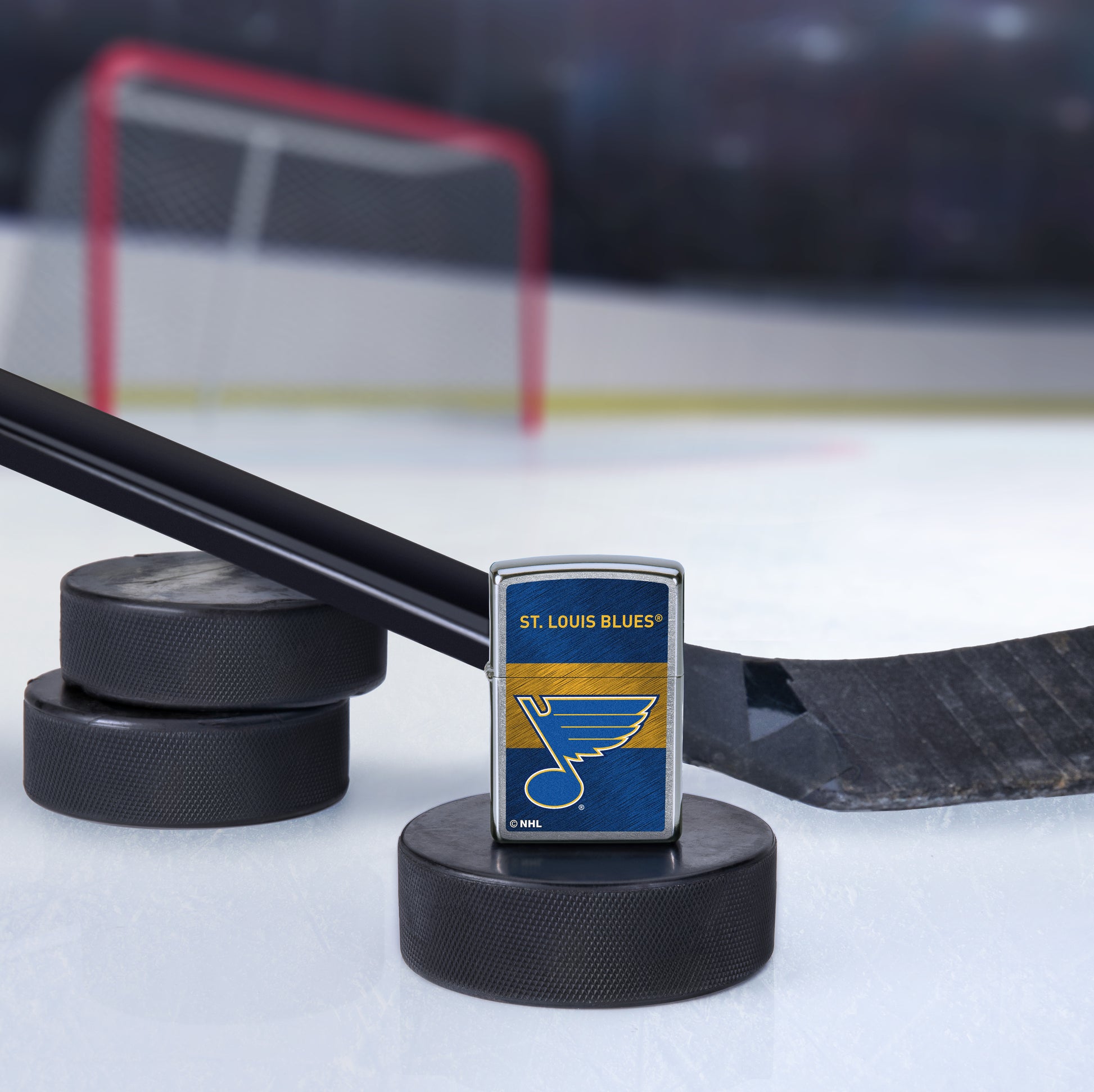 Lifestyle image of the NHL® St Louis Blues™ Street Chrome™ Windproof Lighter standing with a hockey puck and hockey stick, with a hockey net in the background.