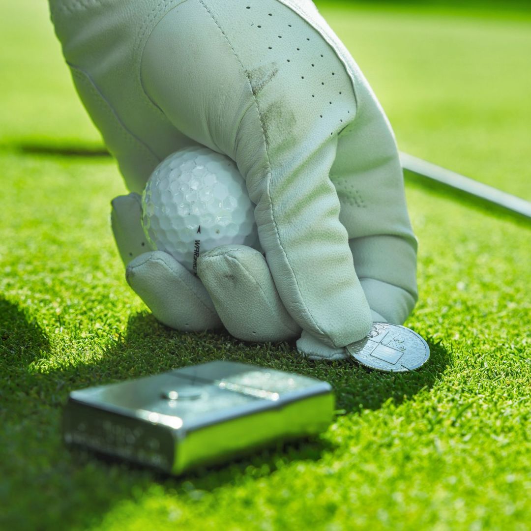 Lifestyle image of Zippo Golf Ball Marker Emblem – Lighter Design Street Chrome Windproof Lighter lying flat on the golf course showing the front, bottom stamp and non-hinge side of the lighter while the golf ball marker is being placed on the green as a ball marker. 

