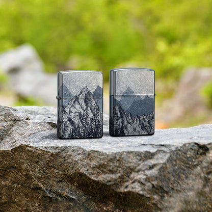 Lifestyle image of two Zippo Mountain Sketch Design 540 Tumbled Chrome Windproof Lighters, one showing the front of the lighter and the other showing the back, standing on a rock with blurred greenery behind them.