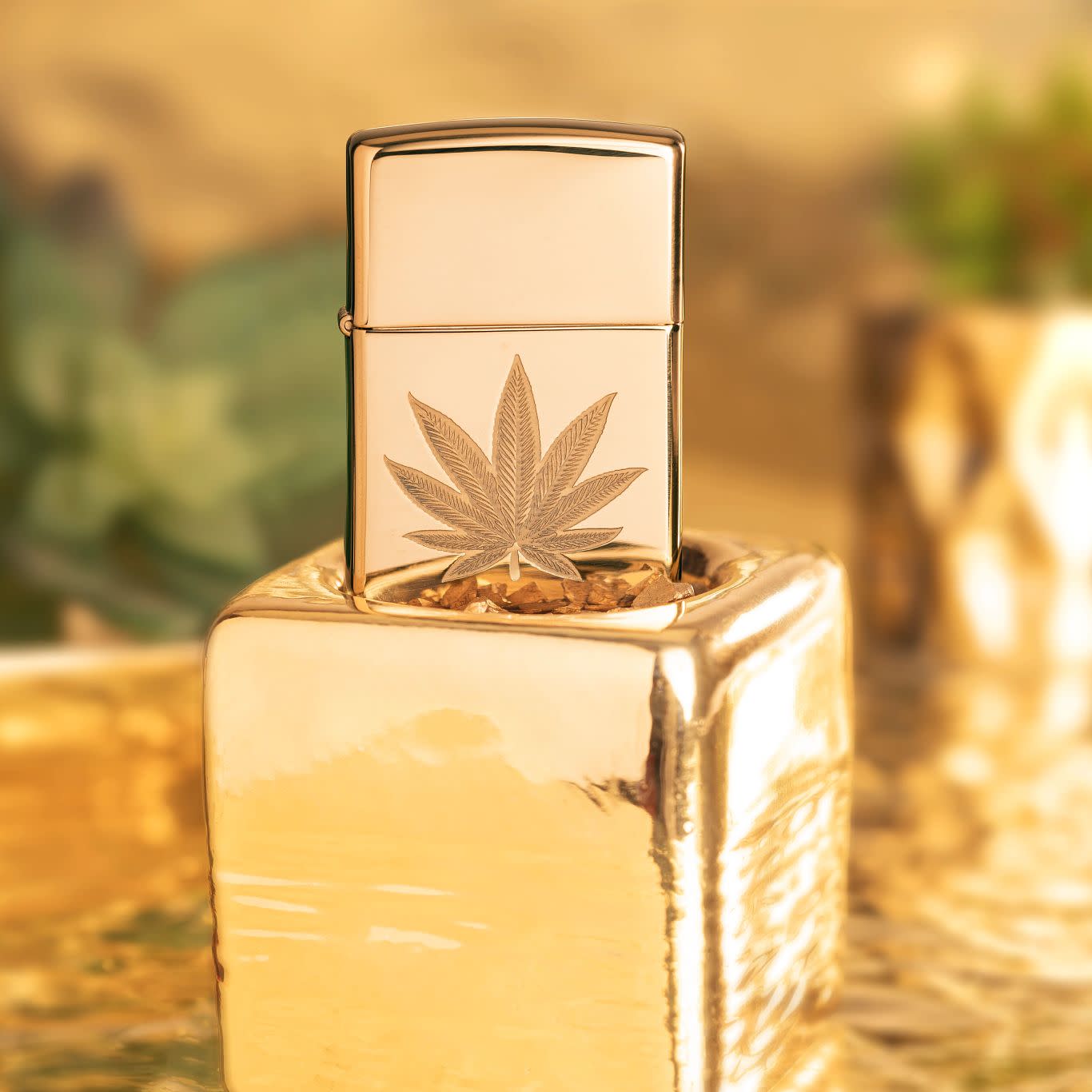 Lifestyle image of Zippo Cannabis Leaf Design High Polish Brass Windproof Lighter standing on a gold cube with succulents in the background.