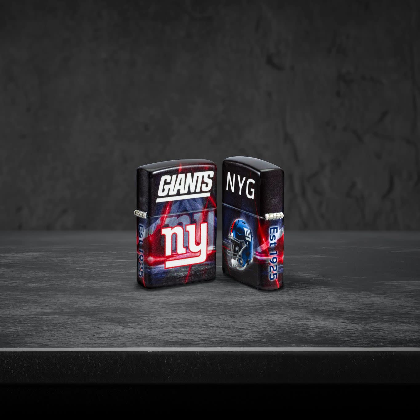 Lifestyle image of two Zippo NFL New York Giants 540 Matte Windproof Lighters, one showing the front of the lighter and the other showing the back, standing on a dark grey surface.