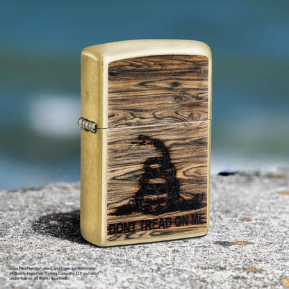 Lifestyle image of Zippo Don't Tread On Me® Woodburn Design Street Brass Windproof Lighter standing on concrete showing the front and hinge side of the lighter with a blurred body of water as the background. 

