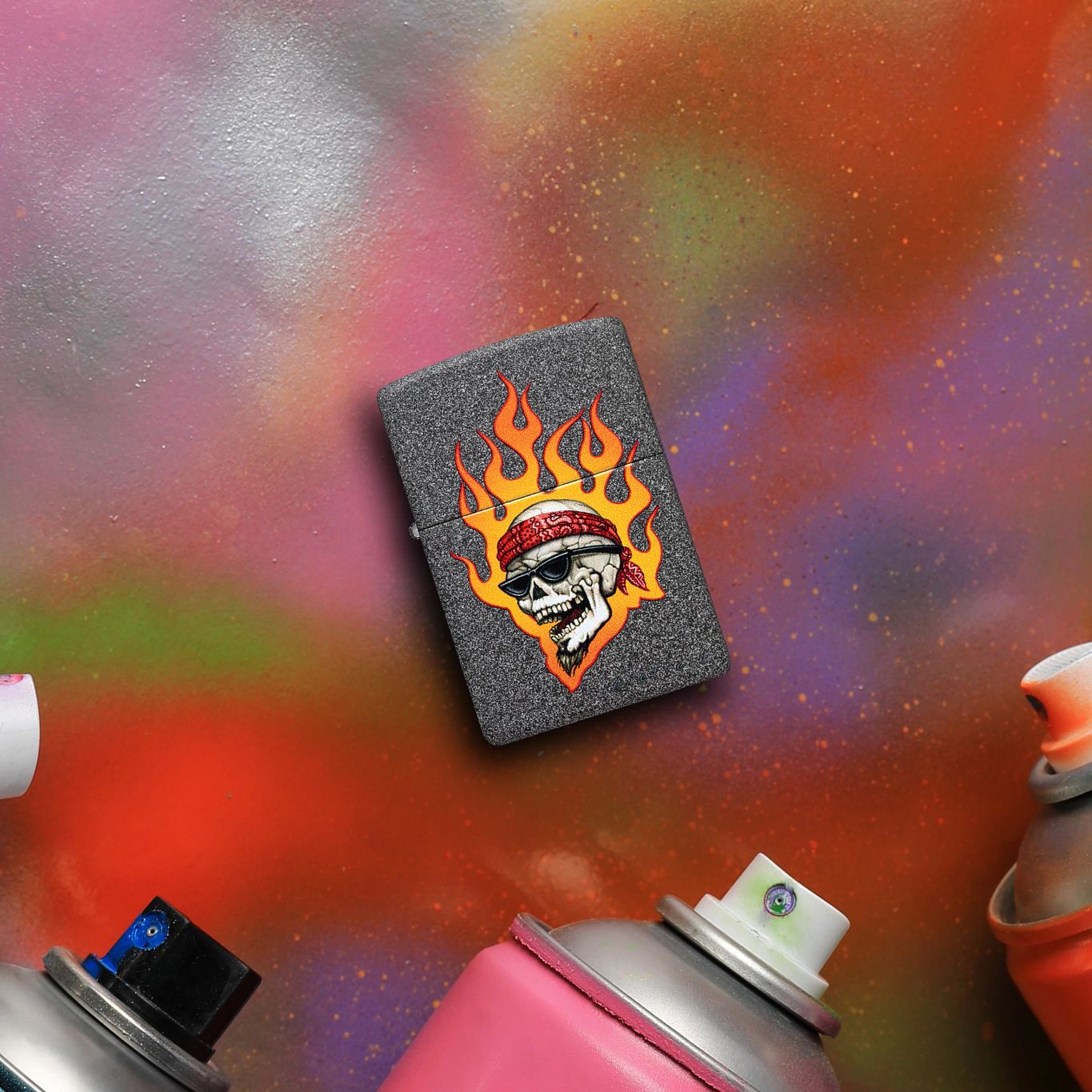 Lifestyle image of Zippo Rietveld Flaming Skull Design Iron Stone Windproof Lighter lying on a graffiti surface next to cans of spray paint while showing the front of the lighter. 


