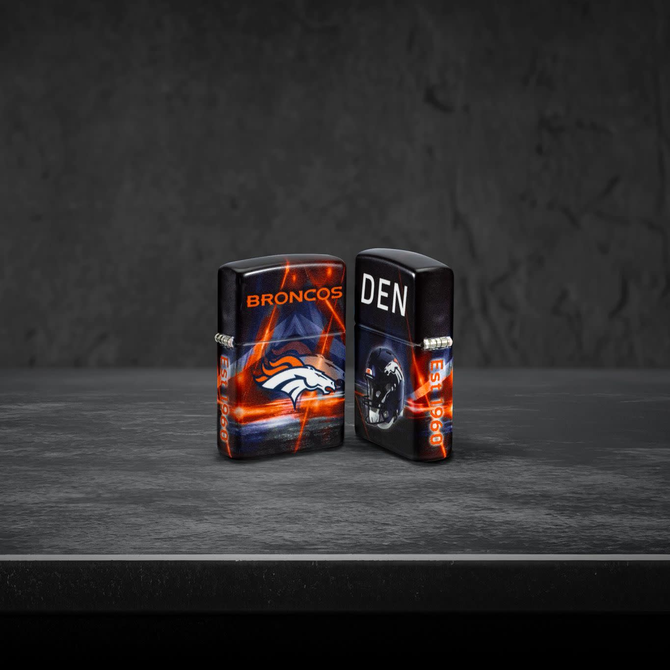 Lifestyle image of two Zippo NFL Denver Broncos 540 Matte Windproof Lighters, one showing the front side of the lighter and the other showing the back, standing on a dark grey surface.