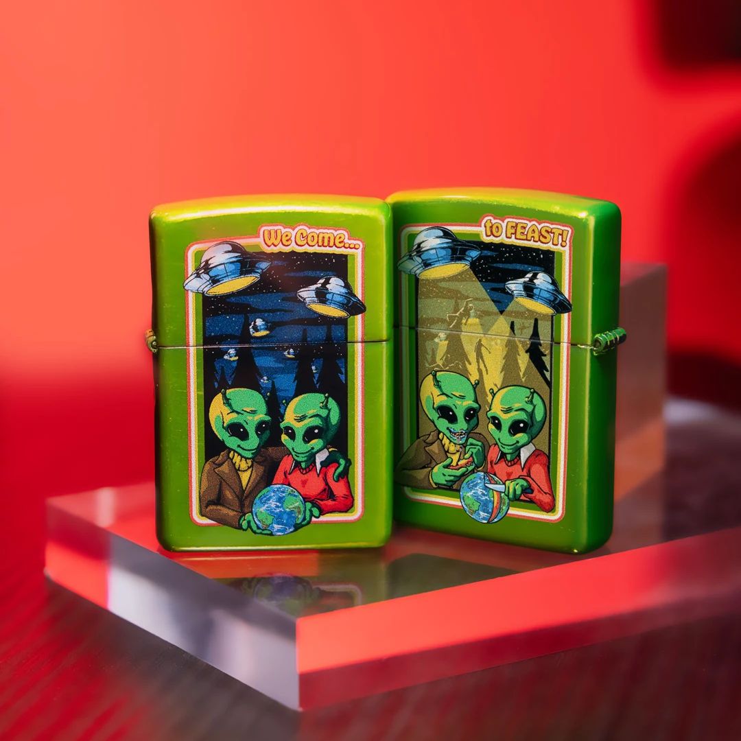 Lifestyle image of two Zippo Take Me To Your Feeder Design Classic Lurid Windproof Lighters; One showing the front of the lighter while the other is showing the back and hinge side of the lighter, both standing on a clear block with a red background and a clear cube behind the lighters. 