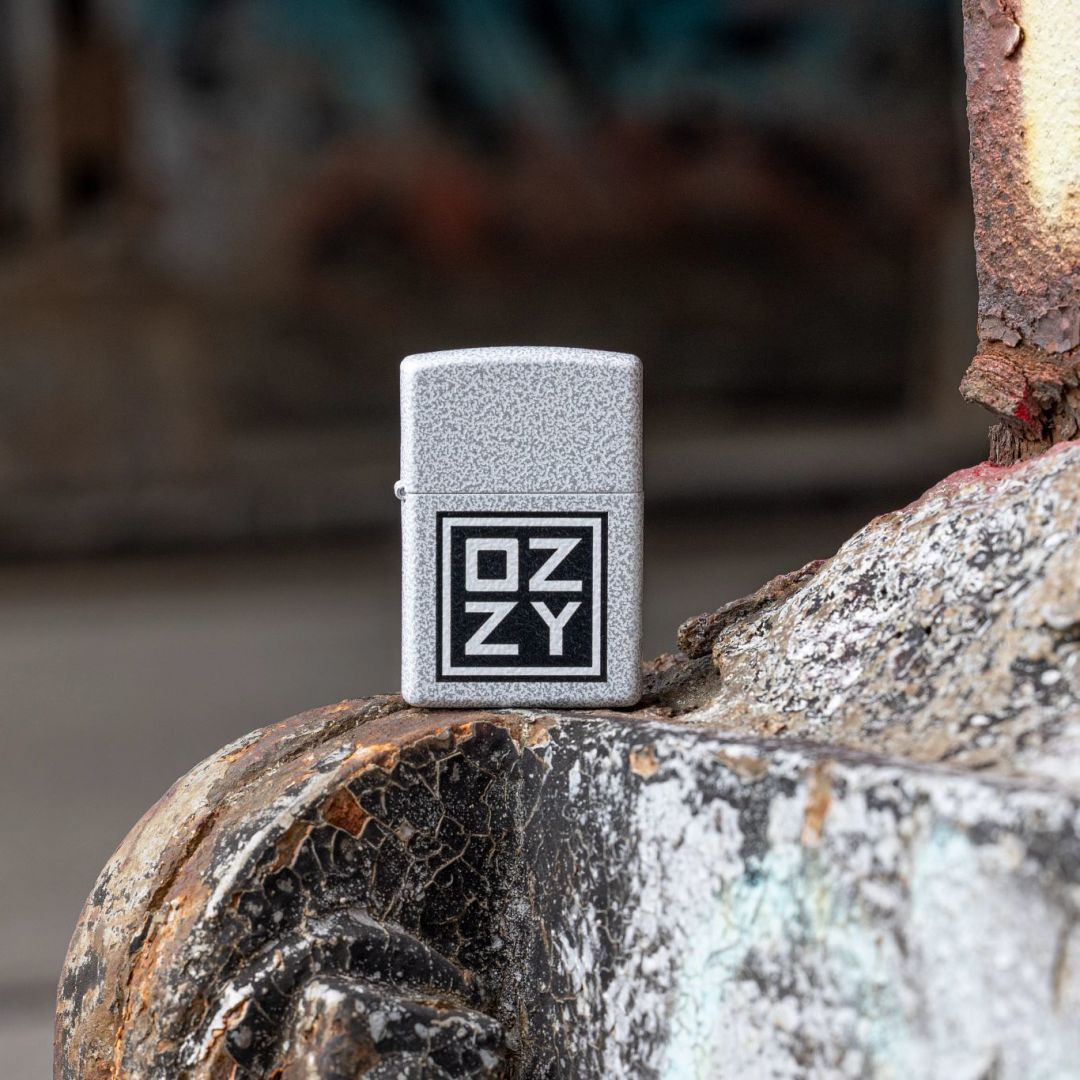 Lifestyle image of Zippo Ozzy Osbourne Block Design Glacier Windproof Lighter standing on a rusty and weathered steel structure showing the front of the lighter with an out of focus background.

