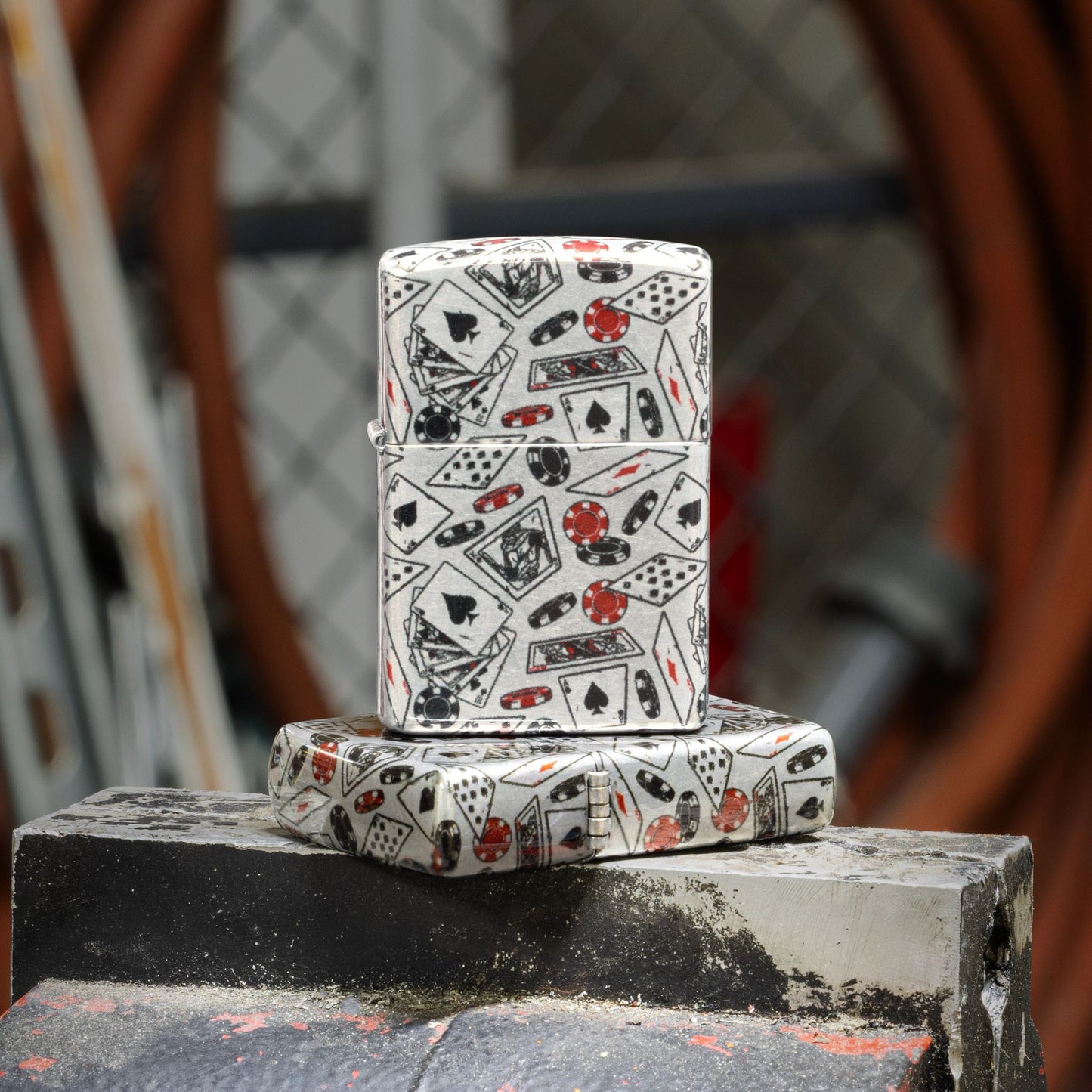 Lifestyle image of two Zippo Let the Chips Fall Design 540 Tumbled Chrome Windproof Lighters; one lying flat on a heavy duty bench vise grip showing the top of the lid and hinge side of the lighter while the other lighter is standing on top of the lighter that is lying flat showing the front of the lighter with a workshop as the background. 