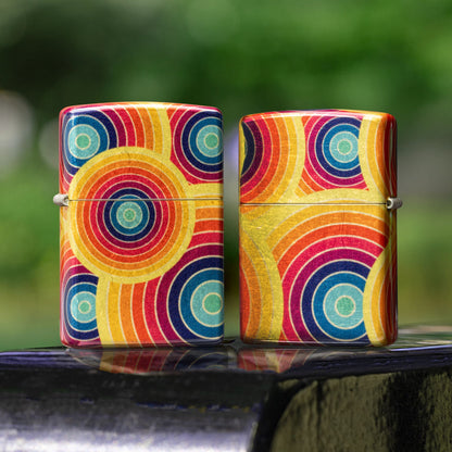 Lifestyle image of two Zippo Funky Fusion Design 540 Tumbled Brass Windproof Lighters; one showing the front of the lighter while the other is showing the back of the lighter, both standing on the armrest of a black metal park bench with an out of focus city park background.

