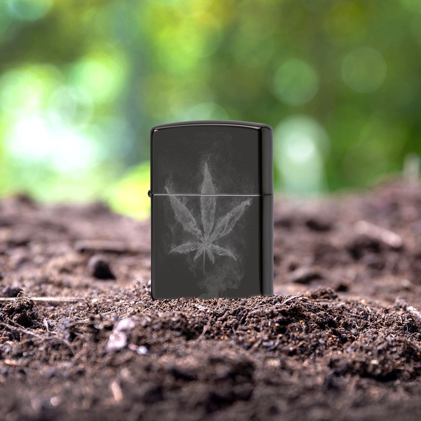 Lifestyle image of Zippo Wispy Leaf Design High Polish Black Windproof Lighter standing in composted foliage with a blurred greenery background. 

