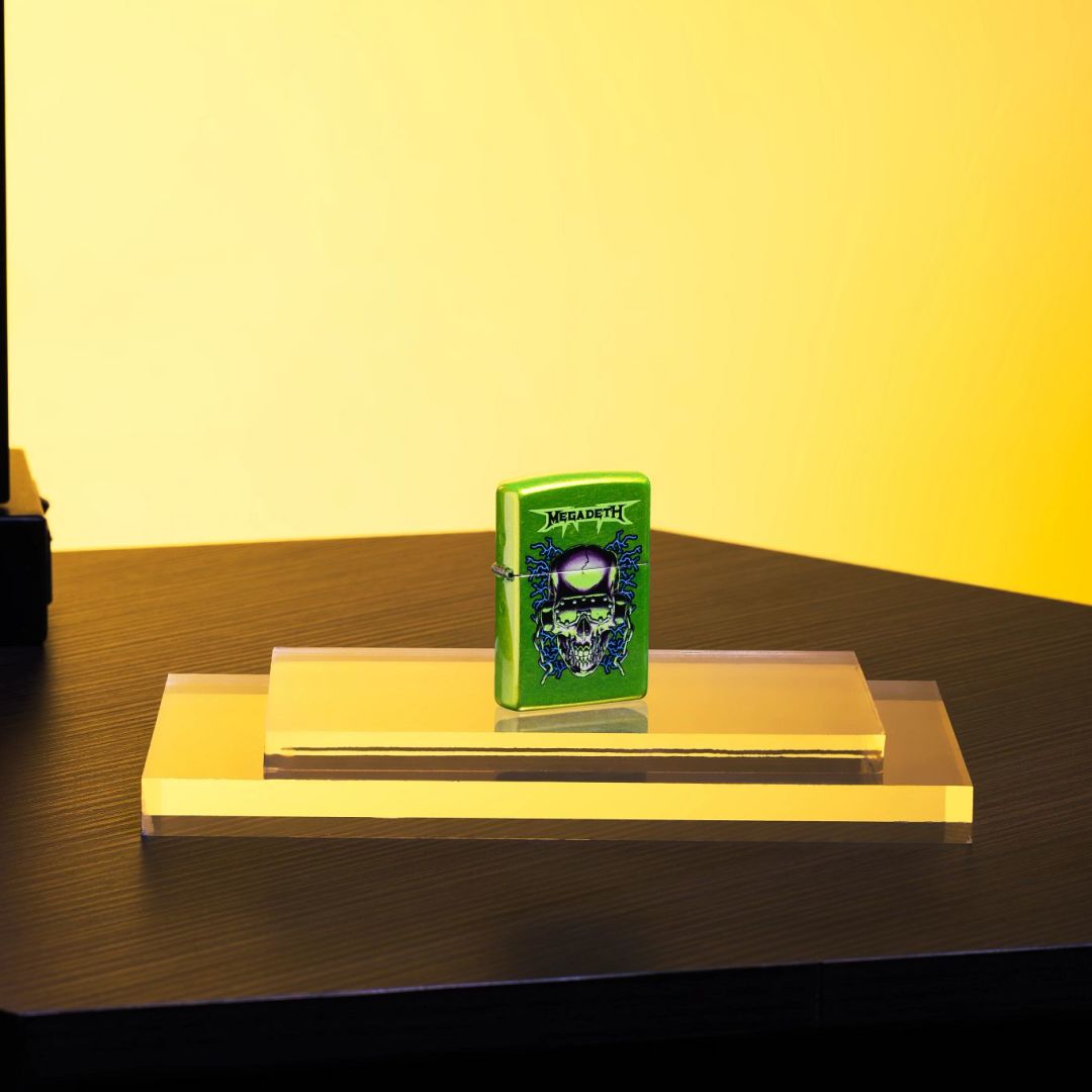 Lifestyle image of Zippo Megadeth Vic Rattlehead Design Classic Lurid Windproof Lighter standing on a clear stand on a table showing the front of the lighter with a yellow-colored background. 

