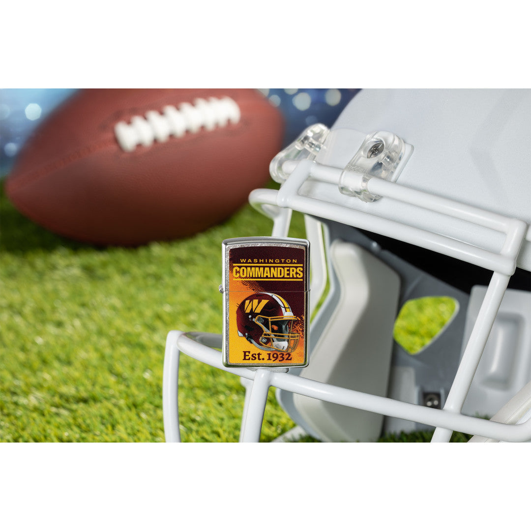Lifestyle image of Zippo NFL Washington Commanders Helmet Design Street Chrome Windproof Lighter standing on the facemask of a helmet showing the front of the lighter with a football lying on a football field as the background.