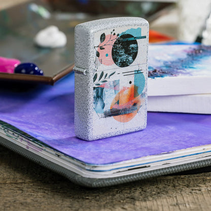 Lifestyle image of Zippo Abstract Collage Design Glacier Windproof Lighter standing on a painted canvas with paint supplies in the background.