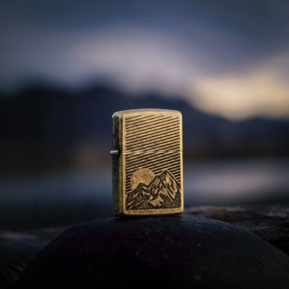 Lifestyle image of Zippo Etched Mountains Design Antique Brass Windproof Lighter standing on a rock showing the front and hinge side of the lighter with an out of focus dark dusk nature background. 

