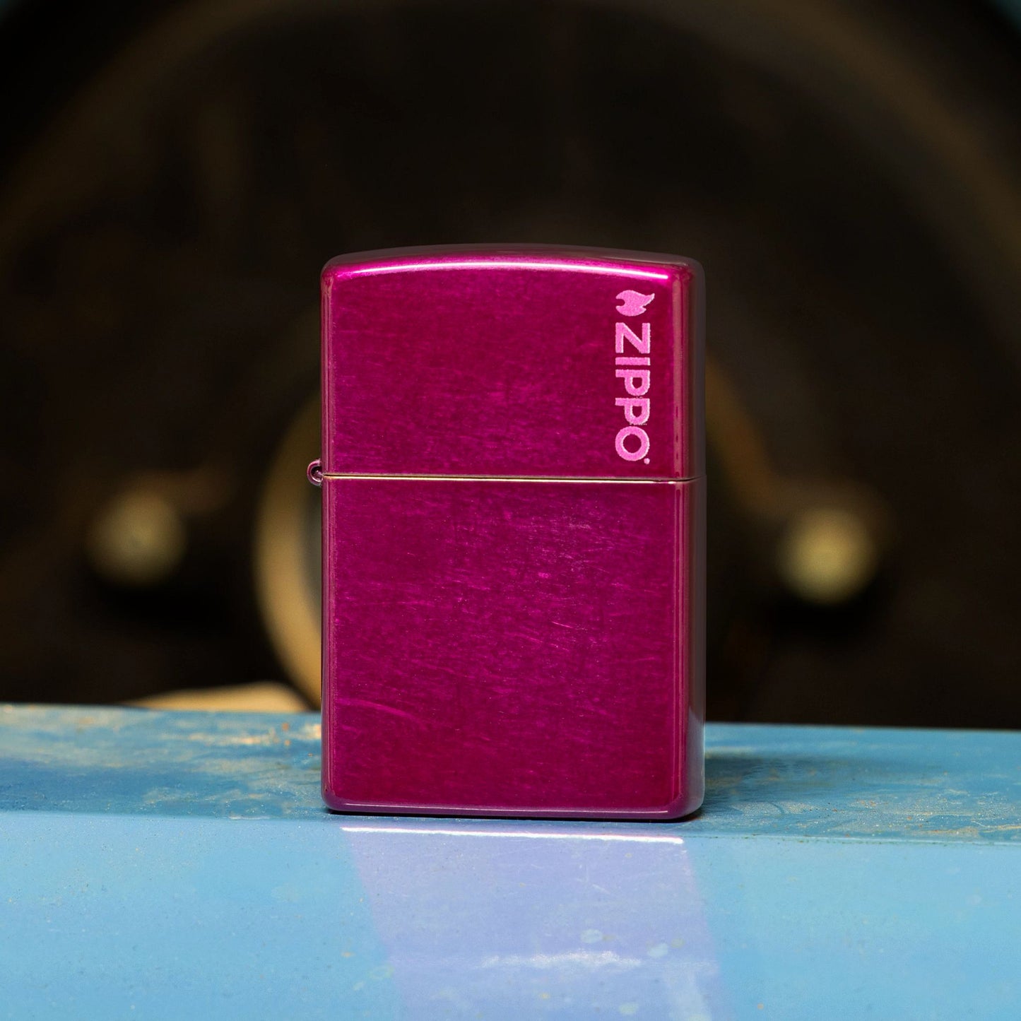 Lifestyle image of Zippo Classic Candy Raspberry Zippo Logo Windproof Lighter standing on a light blue metal bench showing the front of the lighter. 

