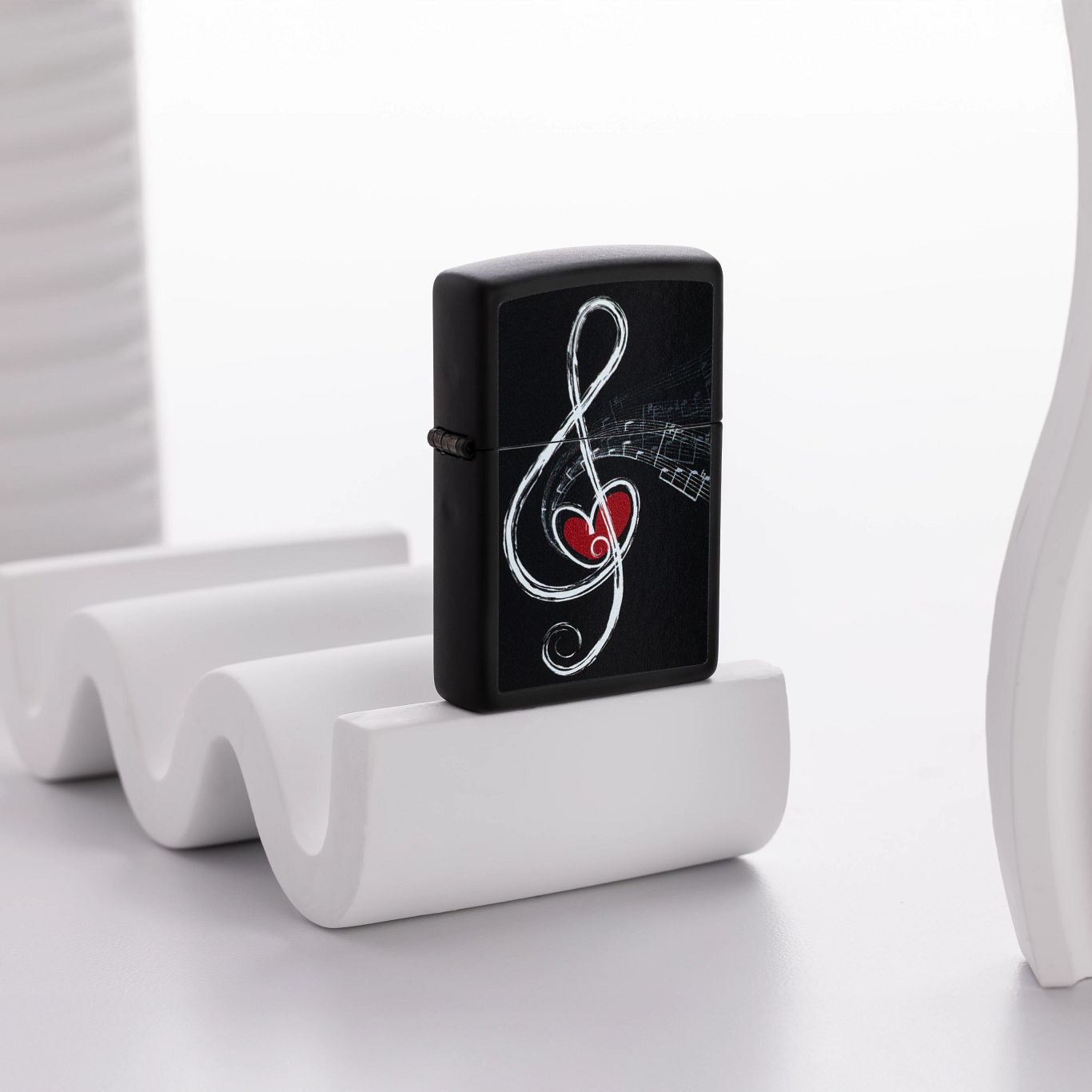 Lifestyle image of Zippo Love Clef Design Black Matte Windproof Lighter standing on a white wavy decorative object next to other white decorative object with a white background. 


