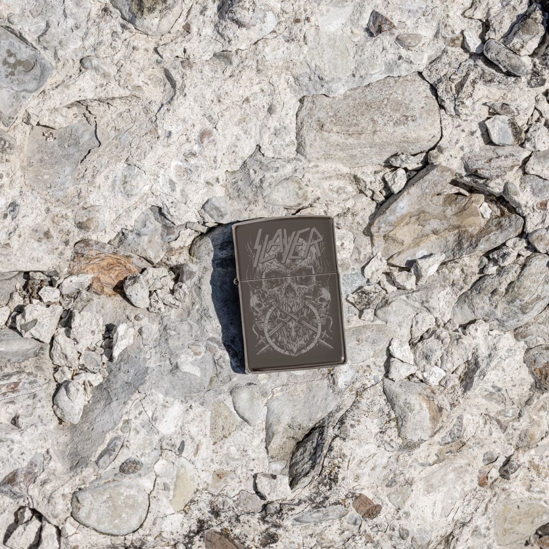 Lifestyle image of Zippo Slayer Skull Design Black Ice® Windproof Lighter lying on a rocky surface showing the front of the lighter. 

