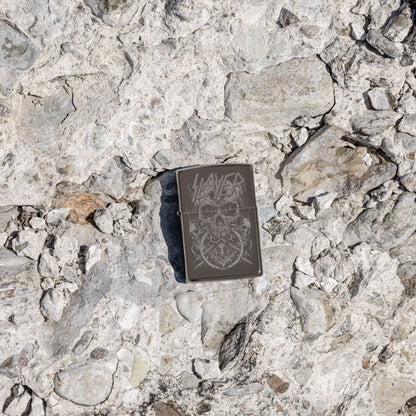 Lifestyle image of Zippo Slayer Skull Design Black Ice® Windproof Lighter lying on a rocky surface showing the front of the lighter. 

