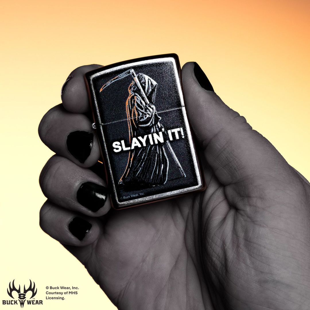 Lifestyle image of Zippo Buck Wear™ Slayin' It Design Street Chrome Windproof Lighter in hand showing the front of the lighter with an orange to yellow ombre background.

