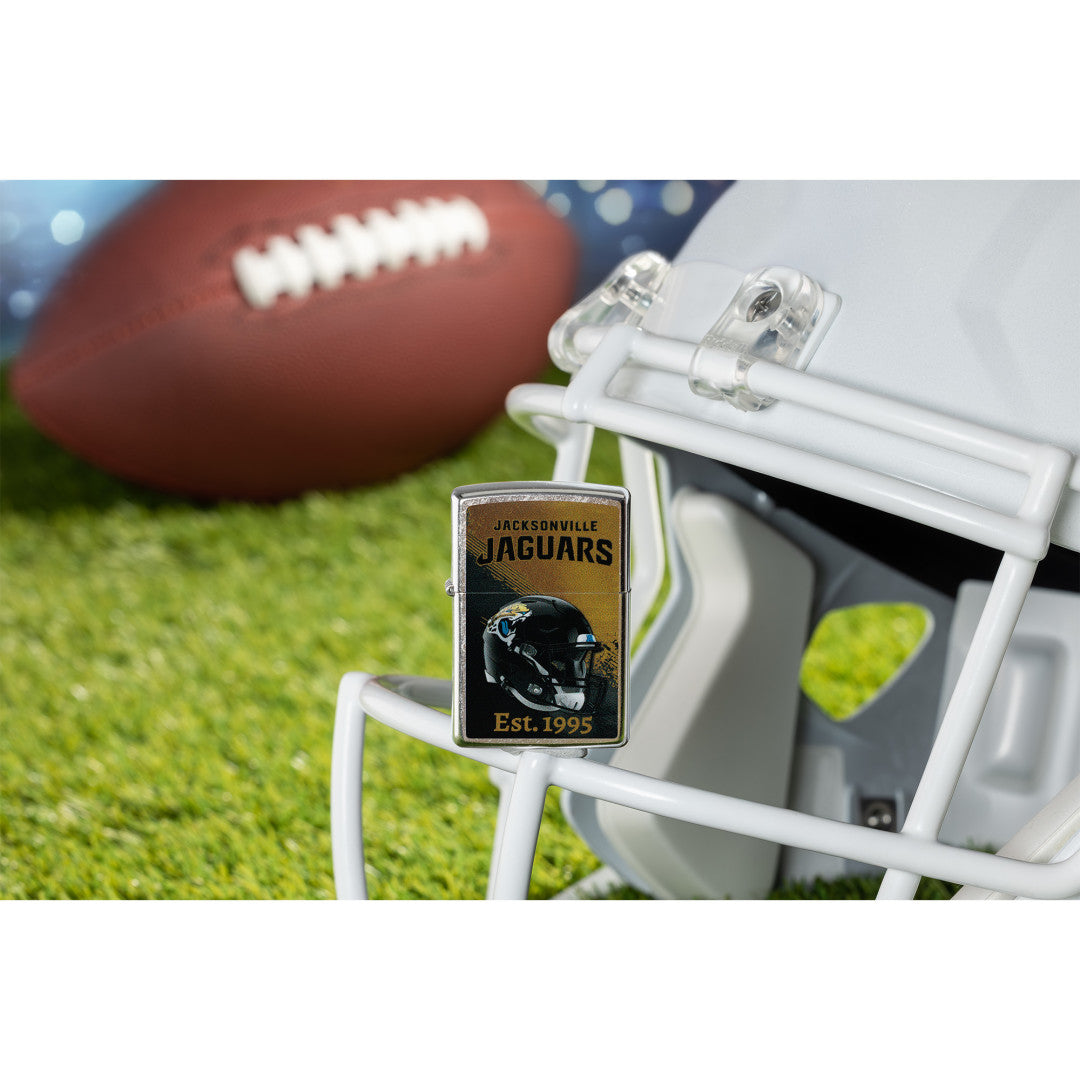 Lifestyle image of Zippo NFL Jacksonville Jaguars Helmet Design Street Chrome Windproof Lighter standing on the facemask of a white helmet showing the front of the lighter with a football lying on a football field as the background.
