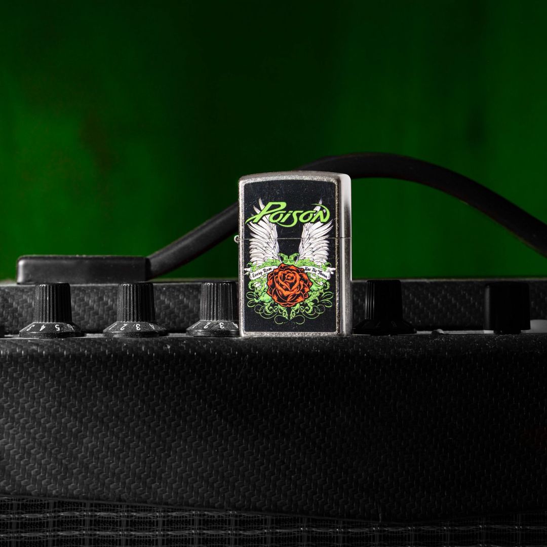 Lifestyle image of Zippo Poison Winged Rose Design Street Chrome Windproof Lighter standing on a black guitar amp showing the front of the lighter with a green background.

