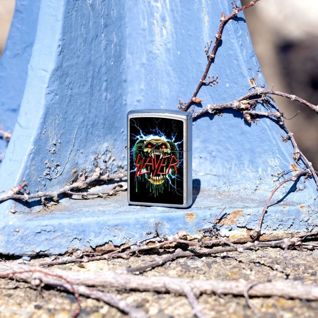 Lifestyle image of Zippo Slayer Snake Design Street Chrome Windproof Lighter standing on a steel surface that is painted light blue showing the front of the lighter with the painted steel and twigs as the background. 


