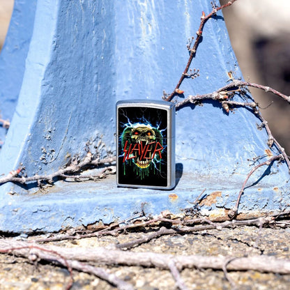 Lifestyle image of Zippo Slayer Snake Design Street Chrome Windproof Lighter standing on a steel surface that is painted light blue showing the front of the lighter with the painted steel and twigs as the background. 

