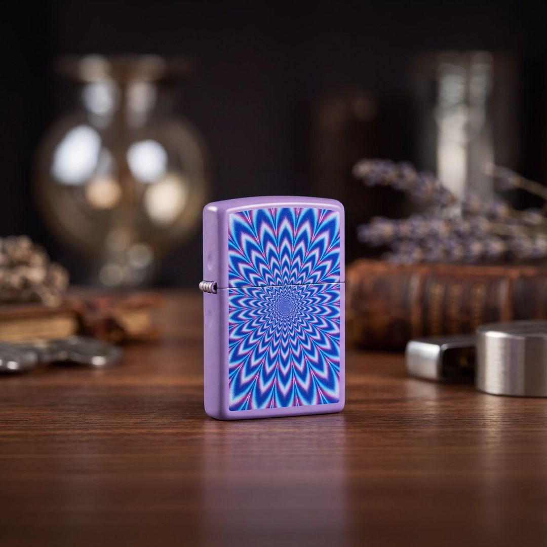 Lifestyle image of Zippo Lavender Illusion Design Smoky Lavender Windproof Lighter standing on a wooden table next to books and table decor showing the front and hinge side of the lighter with an out of focus background.

