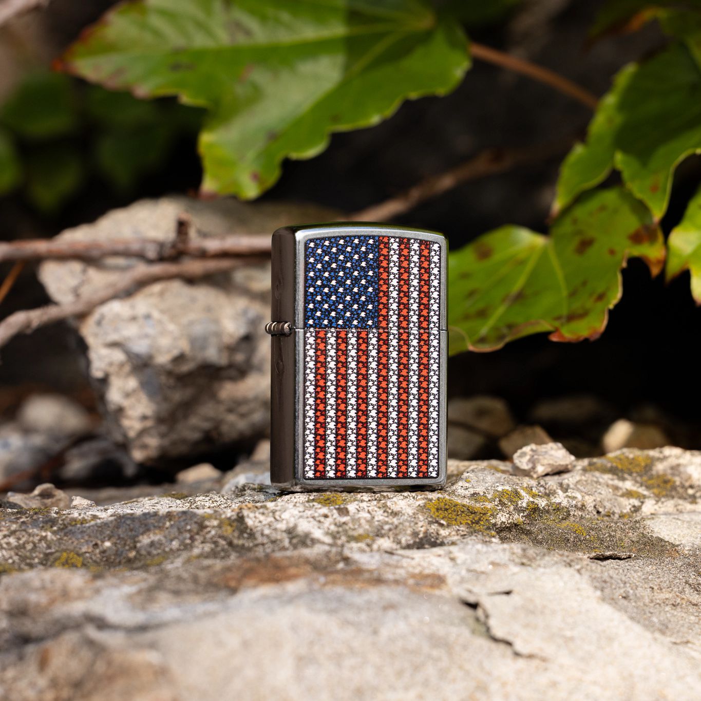 Lifestyle image of Zippo Flame Spangled Banner Design Street Chrome Windproof Lighter standing on a rocky terrain with tiny patches of moss showing the front and hinge side of the lighter with rocks, leaves, and branches in the background. 

