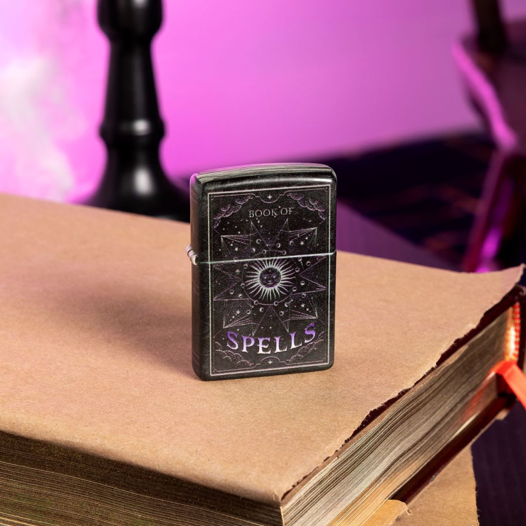 Lifestyle image of Zippo Mystic Book Design 540 Color Glow in the Dark Windproof Lighter standing on a book covered in light brown paper showing the front of the lighter. The background is light purple with the base of a black candelabra behind the lighter. 

