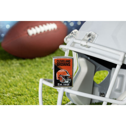 Lifestyle image of Zippo NFL Cleveland Browns Helmet Design Street Chrome Windproof Lighter standing on the facemask of a white helmet showing the front of the lighter with a football lying on a football field as the background. 


