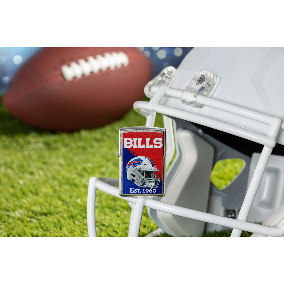Lifestyle image of Zippo NFL Buffalo Bills Helmet Design Street Chrome Windproof Lighter standing on the facemask of a white helmet showing the front of the lighter with a football lying on a football field as the background.

