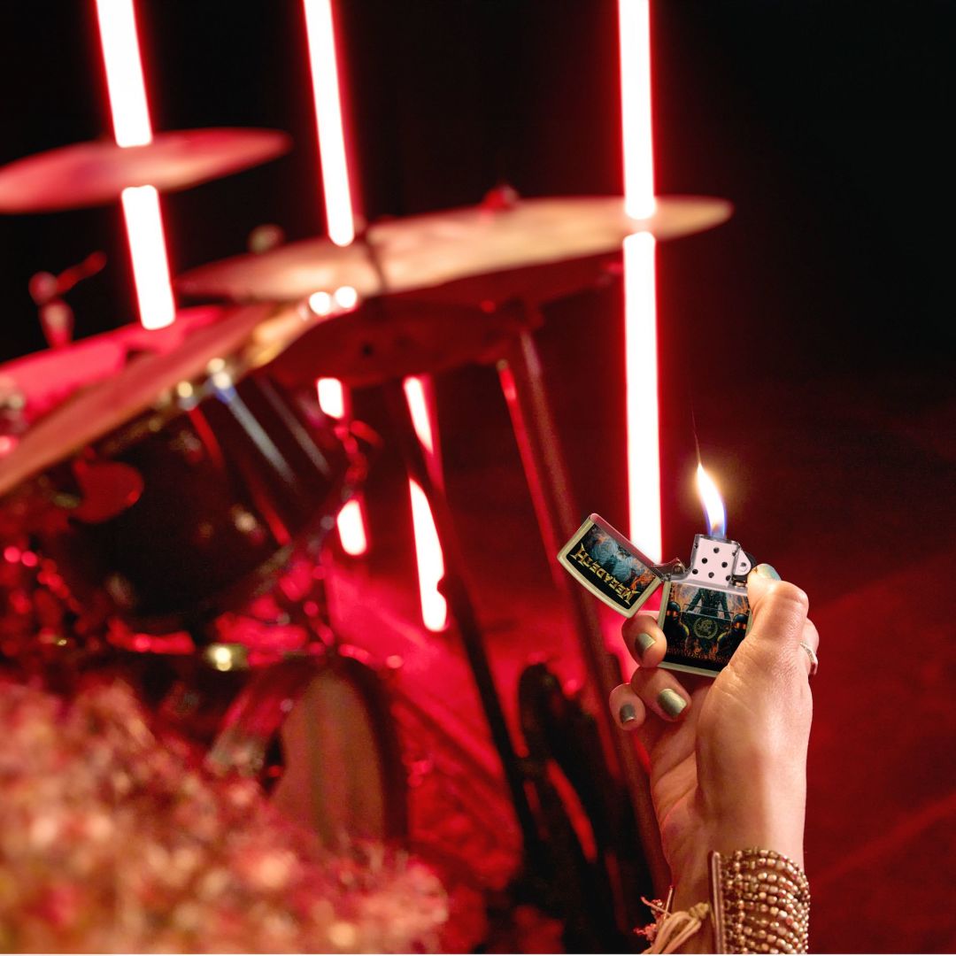 Lifestyle image of Zippo Megadeth New World Order Design Street Brass Windproof Lighter lit in hand showing the front of the lighter with a drum set and red lighting as the background. 

