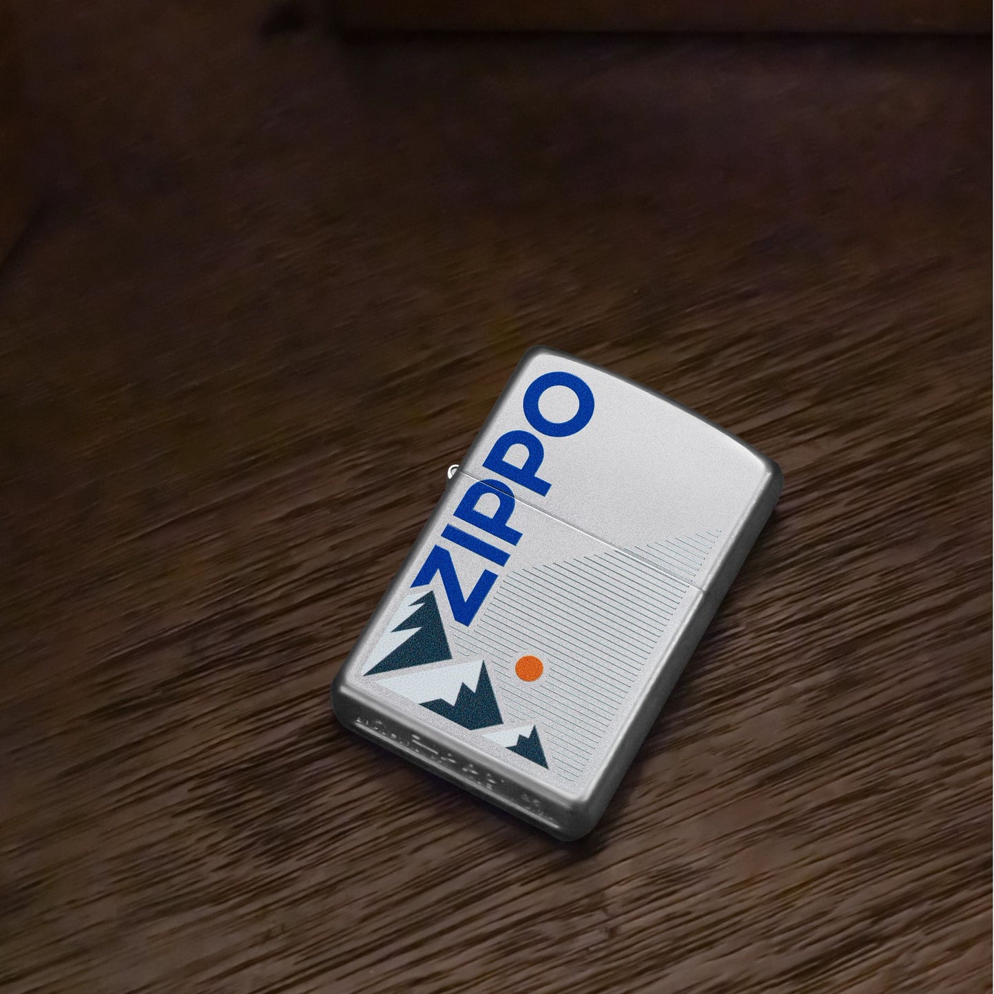 Lifestyle image of Zippo Mountain Design Satin Chrome Windproof Lighter lying flat on a dark wooden table showing the front of the lighter. 

