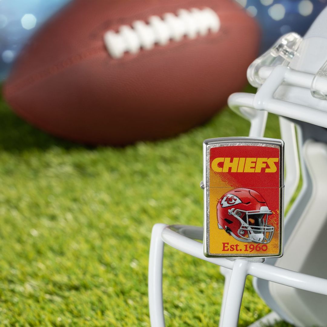 Lifestyle image of Zippo NFL Kansas City Chiefs Helmet Design Street Chrome Windproof Lighter standing on the facemask of a white helmet showing the front of the lighter with a football lying on a football field as the background.