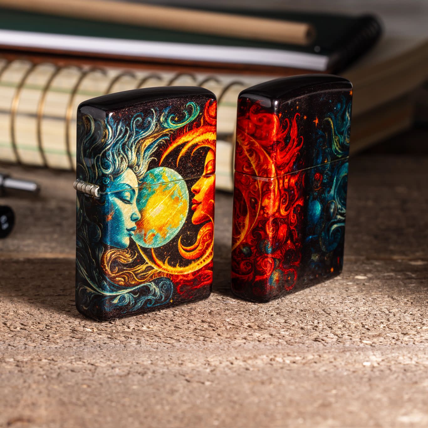 Lifestyle image of two Zippo Sun and Moon Design 540 Tumbled Brass Windproof Lighters standing on a cement surface with pens and a spiral bound notebook in the background.