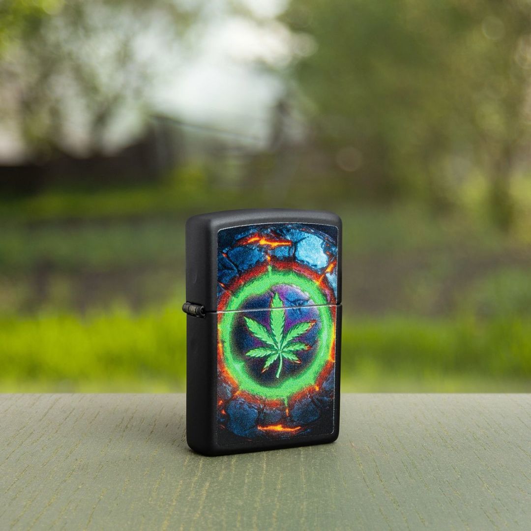 Lifestyle image of Zippo Lava Leaf Design Black Matte Windproof Lighter standing at a 3/4 angle on a green wooden surface showing the front and hinge side of the lighter with an out of focus background with green grass and trees. 

