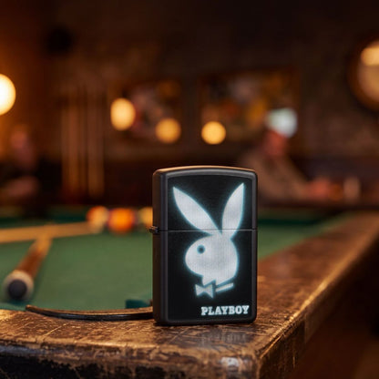 Lifestyle image of Zippo Playboy Blur Design Black Matte Windproof Lighter standing on an old pool table showing the front of the lighter with an out of focus bar as the background. 

