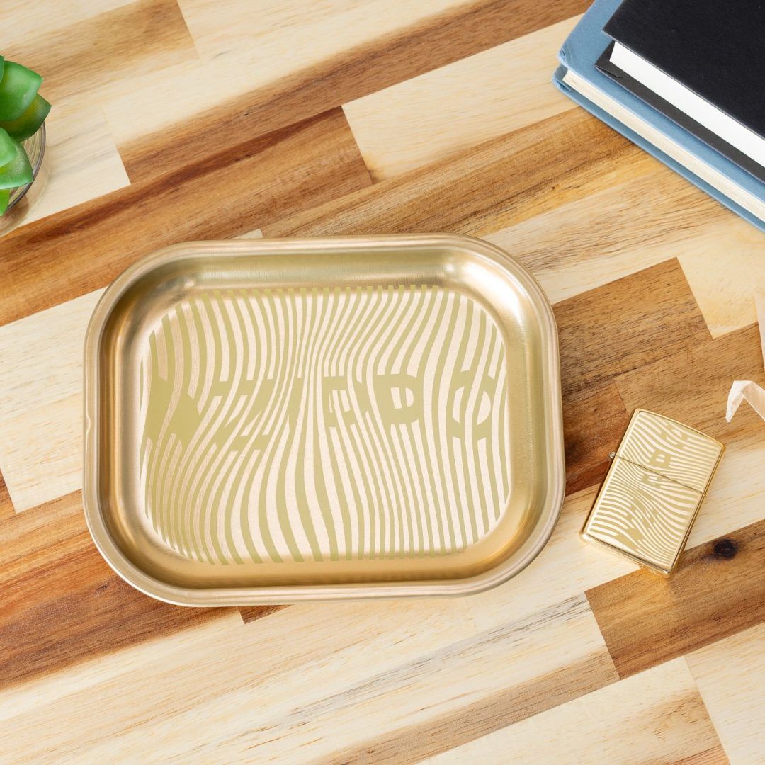 Lifestyle image of Rolling Tray and Lighter Gift Set lying on a two-toned wooden table next to some books, a succulent and an origami crane, showing the front of the lighter and top view of the rolling tray. 

