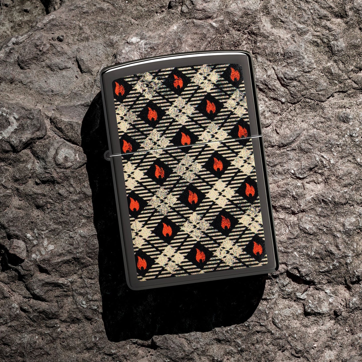 Lifestyle image of Zippo Tartan Design High Polish Black Windproof Lighter lying on a rocky surface while showing the front of the lighter. 

